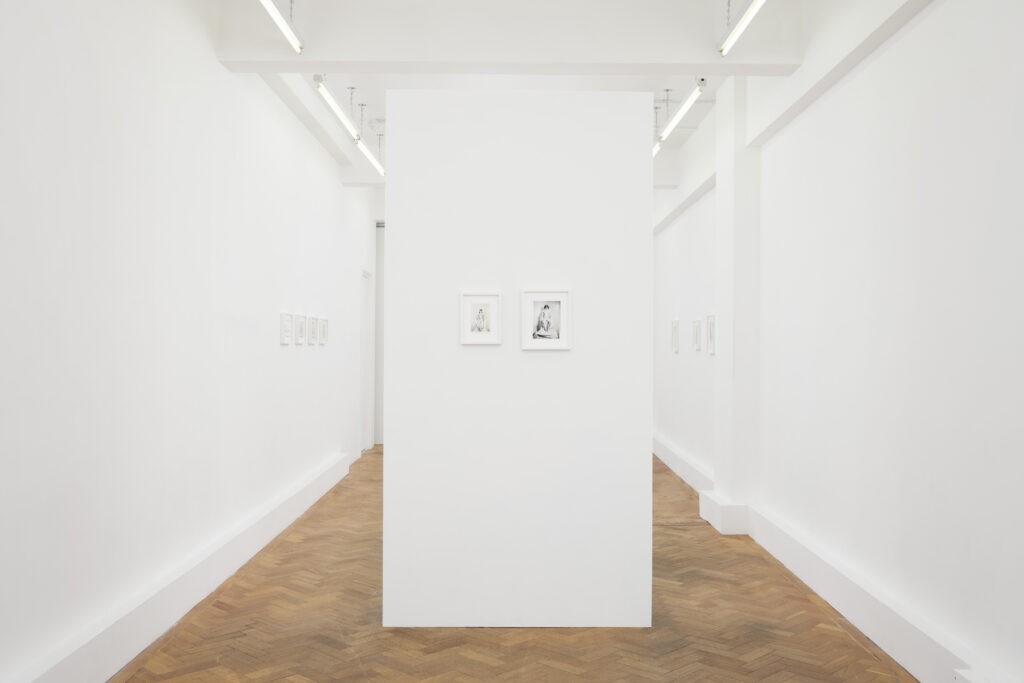 A minimalist art gallery interior featuring a white wall with two framed artworks displayed on it, surrounded by an empty, well-lit space.