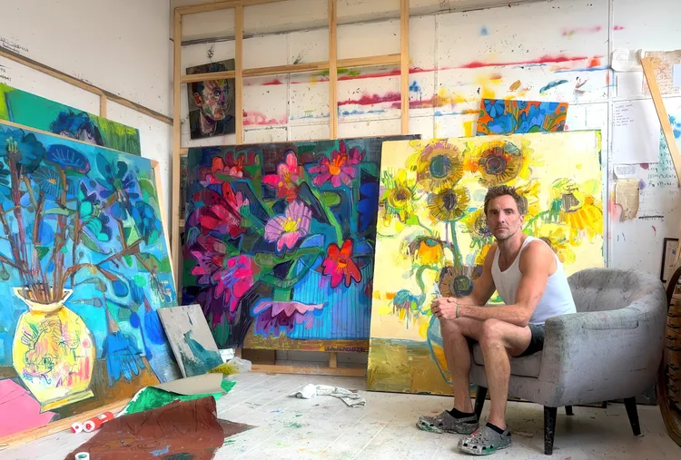 A man sitting in a gray chair in an art studio filled with colorful floral paintings. | 2026 BEERS London