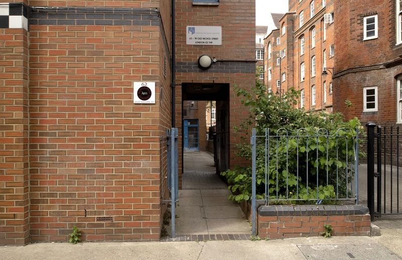 A narrow pathway between buildings with brick walls, featuring a gate and greenery on the side.