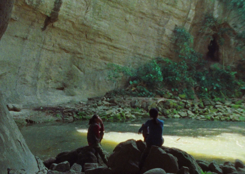 Two people sitting on rocks near a calm body of water surrounded by steep rocky cliffs and greenery.