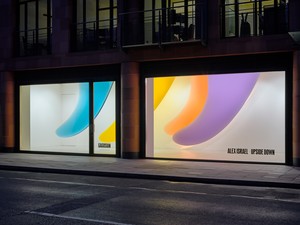 A storefront at night with two large, colorful window displays featuring abstract curved shapes in blue, yellow, and purple.