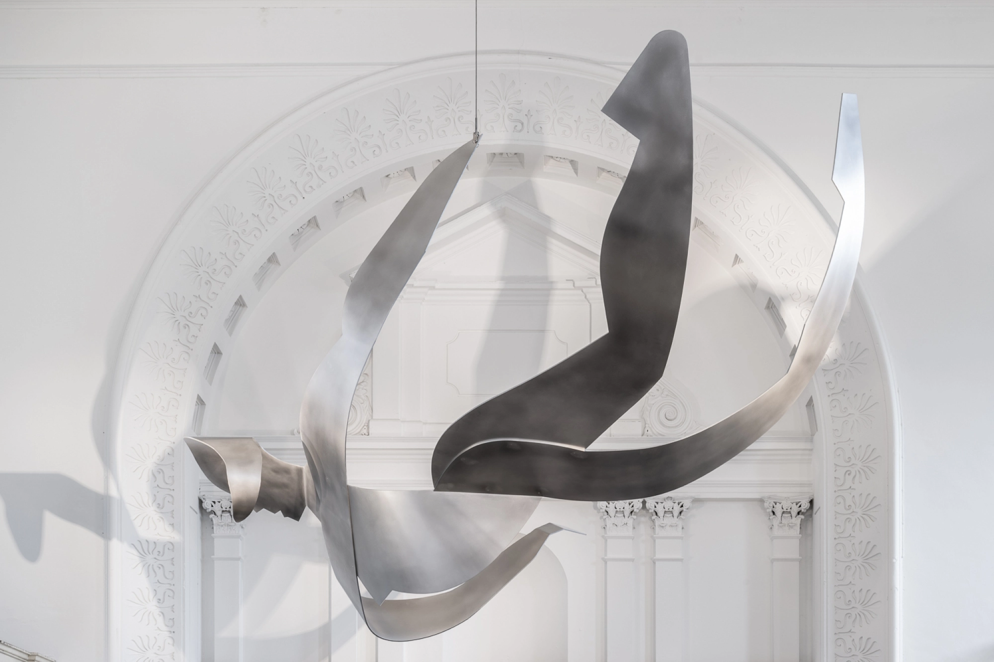 A large, abstract metal sculpture hanging in a bright, minimalist space with white walls and architectural details.