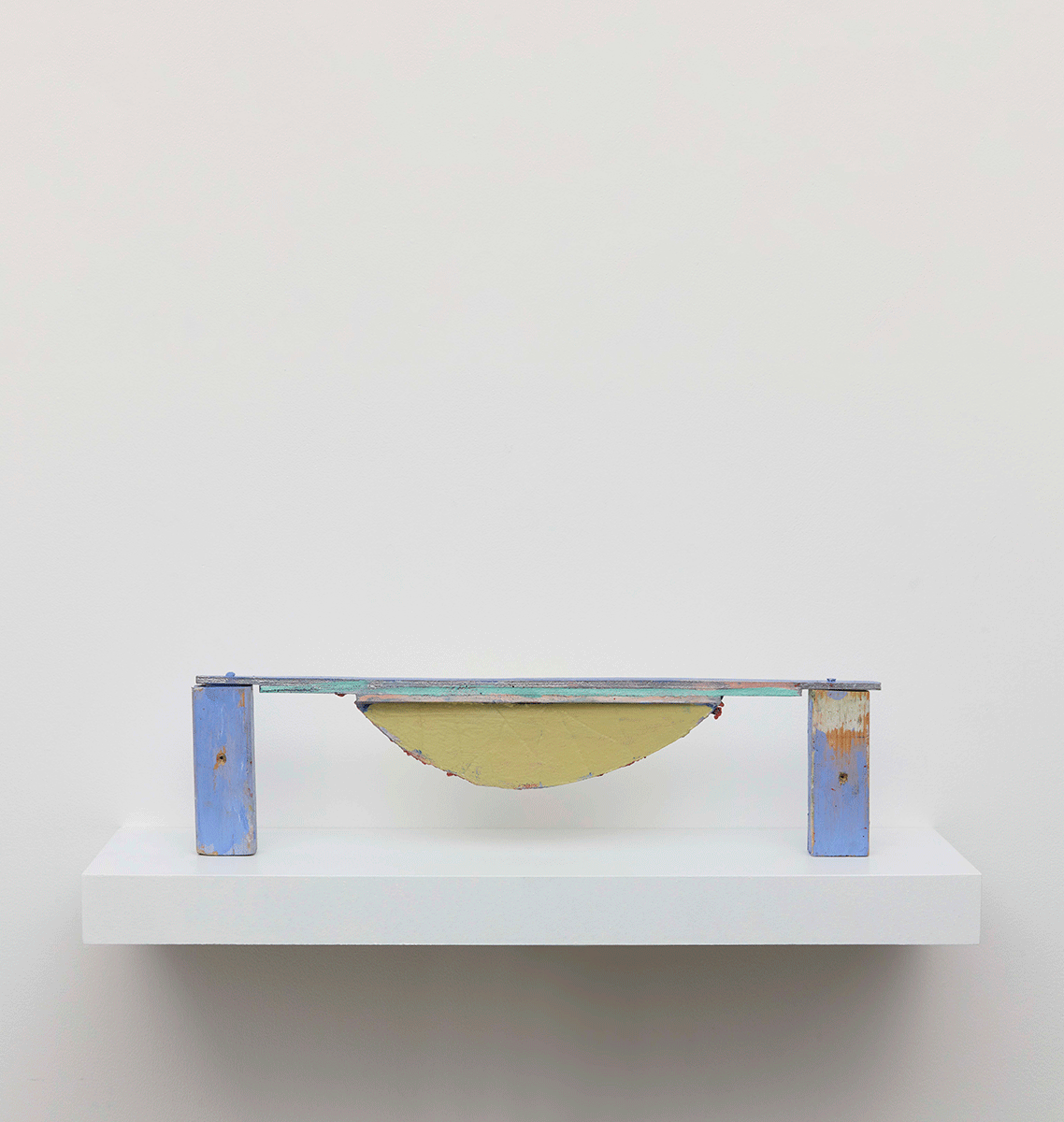 A minimalist sculpture displayed on a white shelf with a pale yellow curved form supported by two blue metallic supports. The piece shows signs of wear and rust.
