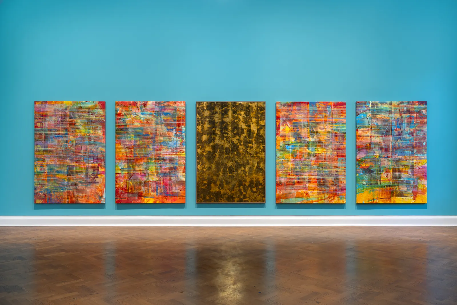 Five abstract paintings on a bright blue wall in an art gallery. The paintings feature a variety of colors and patterns, with one predominantly gold piece among them.
