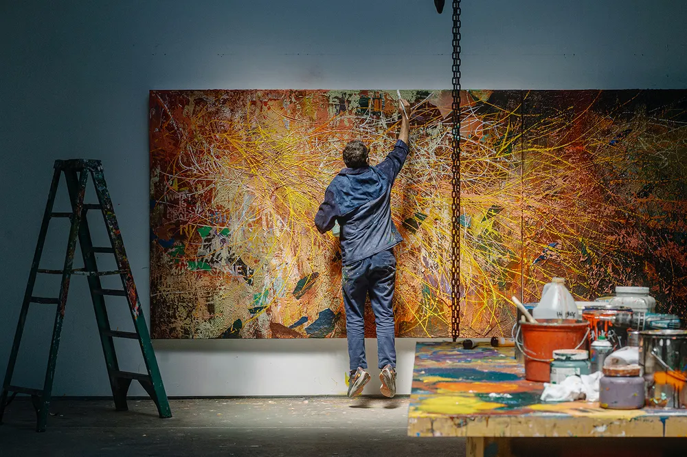 A person creating art on a large canvas in a studio, with paint supplies and a ladder visible.