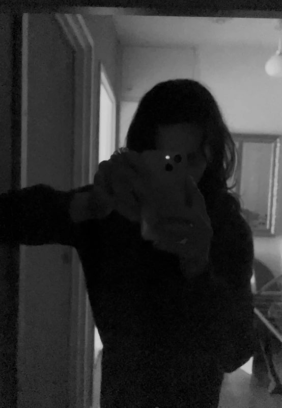 A person taking a selfie in a dimly lit room, holding a smartphone and standing in front of a mirror.