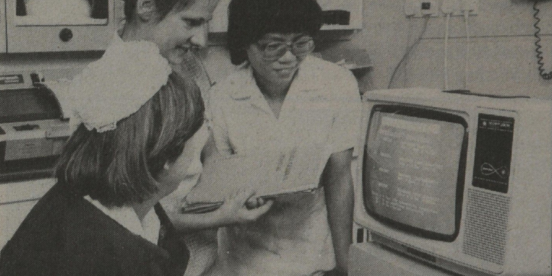 Three individuals, two women in nurse uniforms and one woman in a white shirt, are gathered around a computer monitor, engaged in discussion. The image appears to be in black and white.