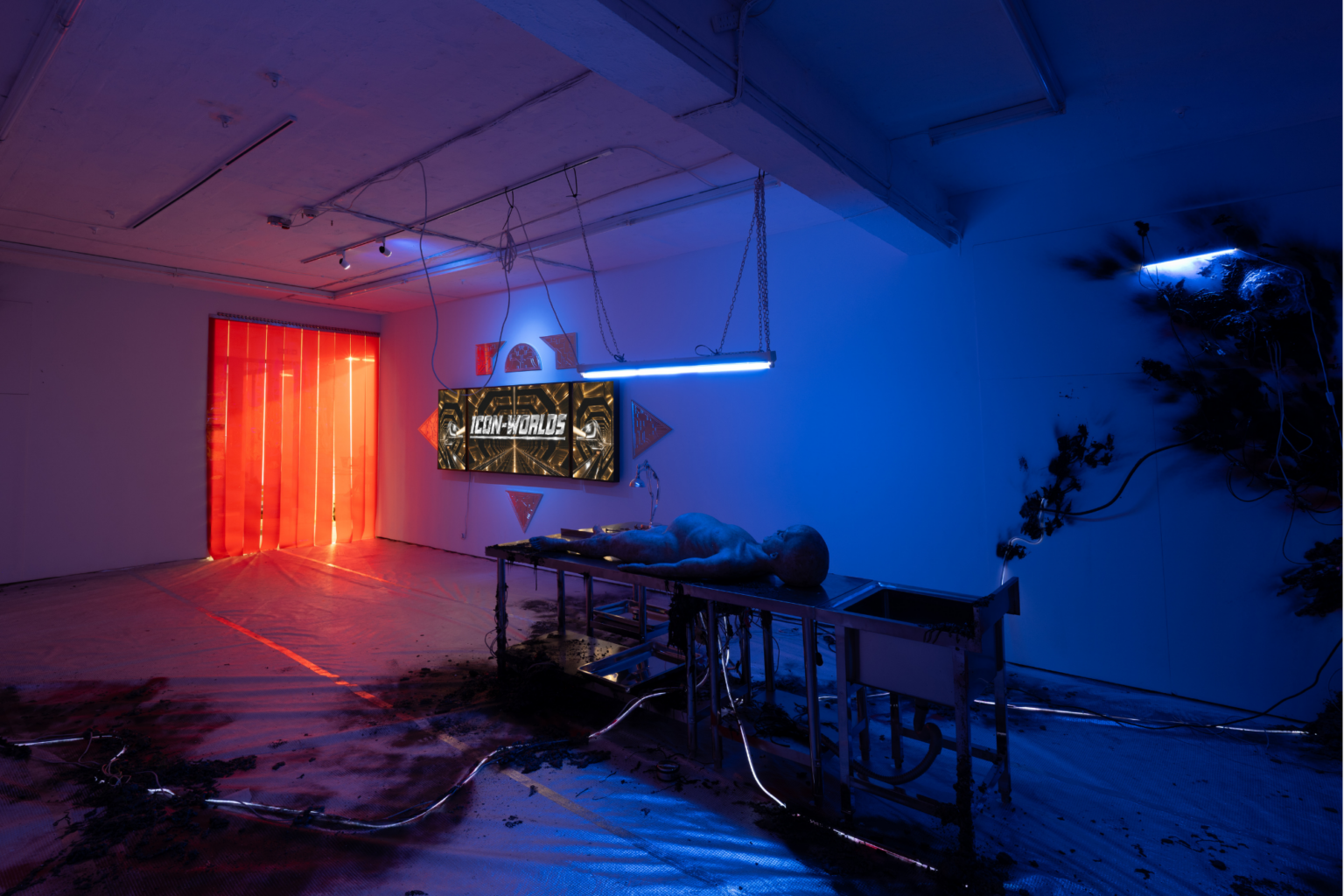 An art installation featuring a dimly lit room with blue and red lighting, a table with a lifelike figure lying on it, and a large screen displaying the text "NEW WORLDS."