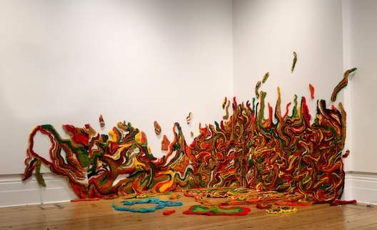 An abstract fiber art installation featuring vibrant, swirling patterns in red, yellow, green, and other colors, resembling flames or waves, extending from the floor to the wall of an art gallery.