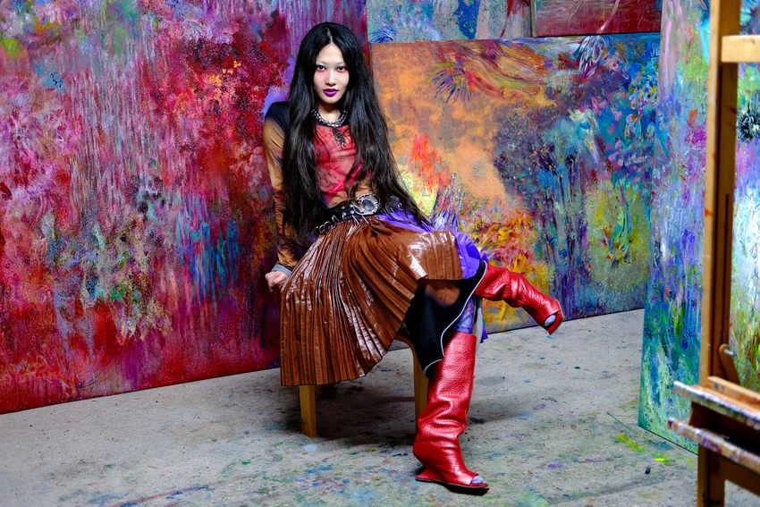 A person with long black hair sits on a wooden chair, surrounded by colorful abstract paintings. They are wearing a red and brown outfit with shiny, knee-high red boots.