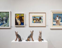 A gallery display featuring animal-themed artwork, including paintings of foxes and ducks, alongside four ceramic fox sculptures arranged on a pedestal.