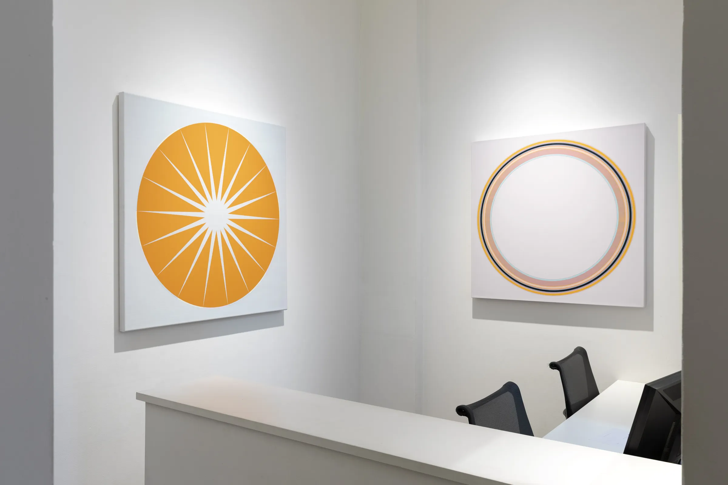 Two abstract paintings hang on the walls of a modern interior. One painting features a bright orange sunburst design, while the other is a circular pattern with a white background and concentric circles in shades of orange and yellow. A reception desk with two black chairs is visible below the artworks.