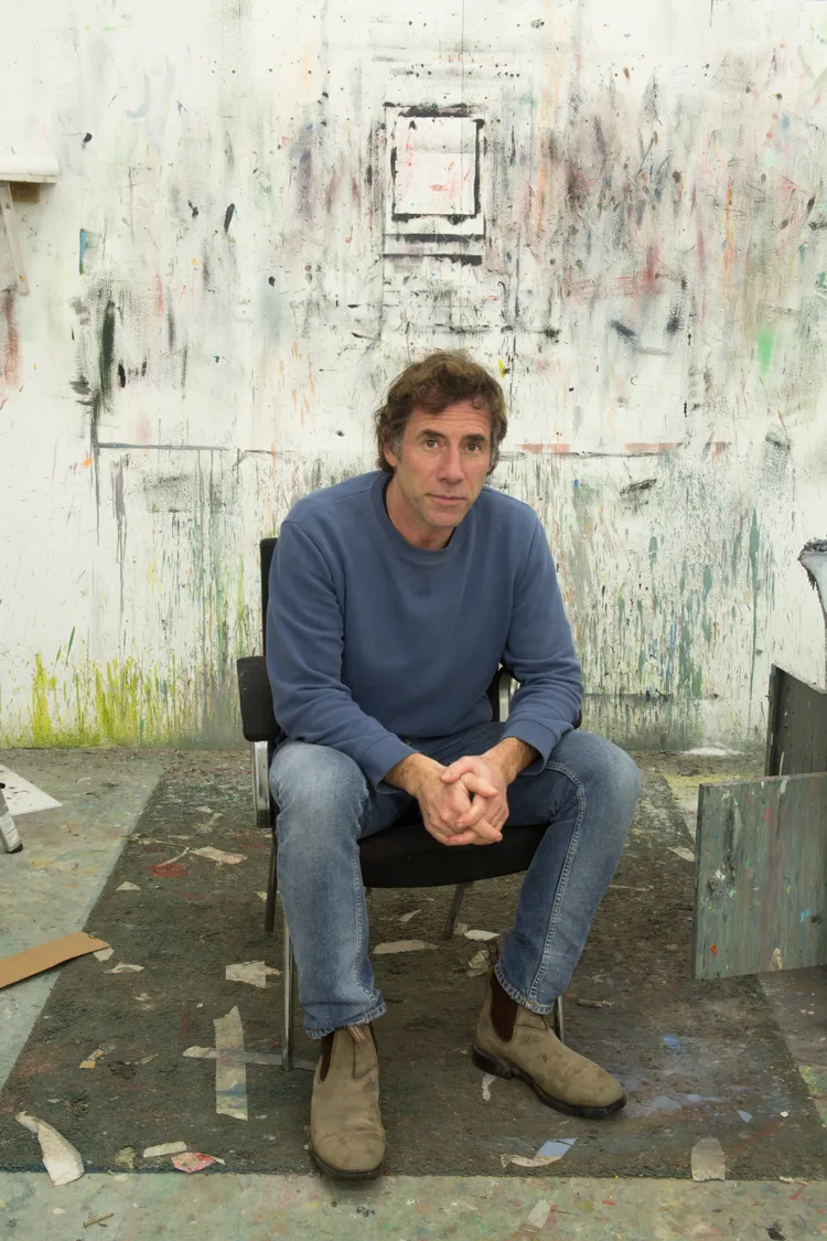 A man sitting on a chair in a messy art studio with splattered paint on the walls and floor.