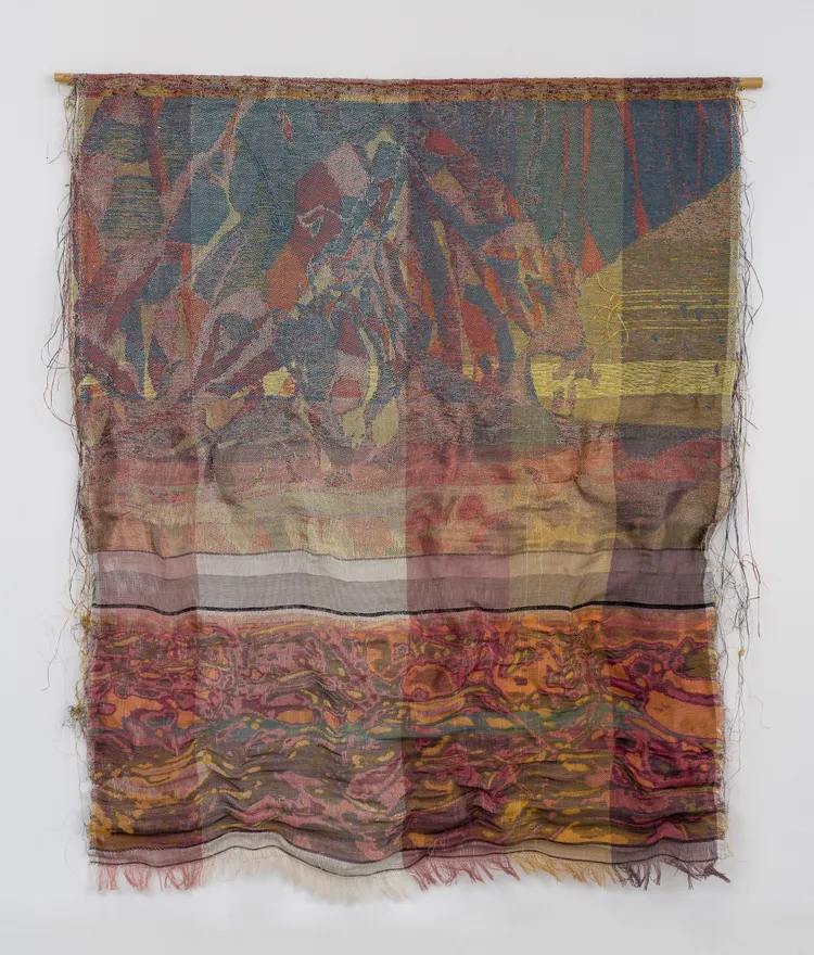 A colorful textile artwork featuring abstract patterns and textures, with varying shades of red, orange, blue, and yellow. The fabric is draped and has loose threads along the bottom edge.