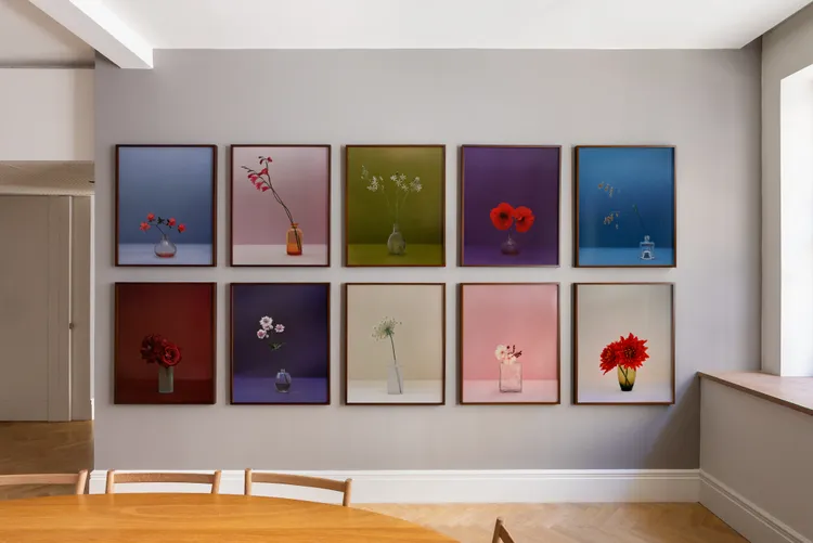 A stylish interior with a wall displaying ten framed floral photographs in various colors, alongside a light wooden dining table and minimalist decor.