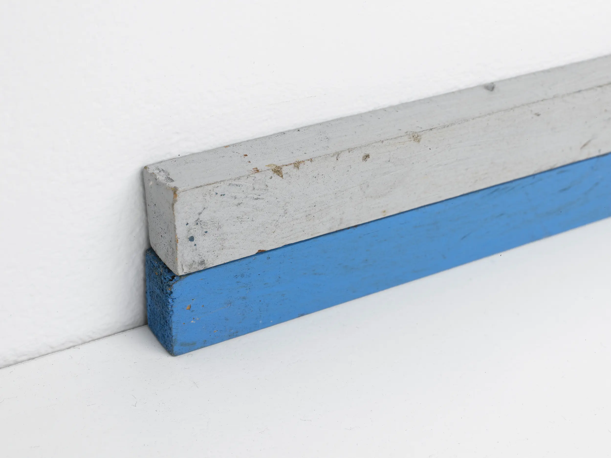 A gray and a blue wooden plank resting against a white wall.