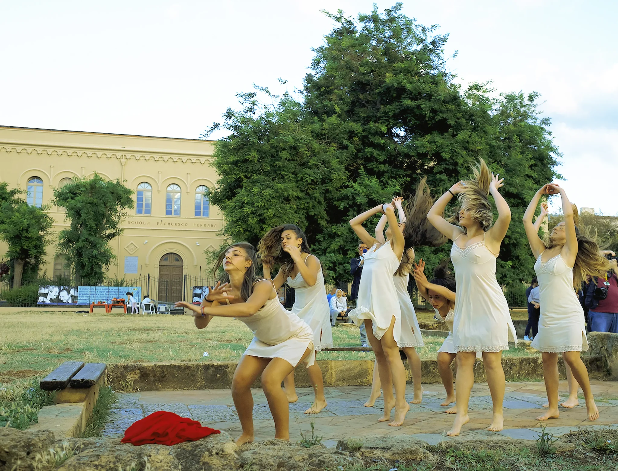 A group of dancers in white dresses performing outside, with some jumping and others posing, in a grassy area with trees and a building in the background.