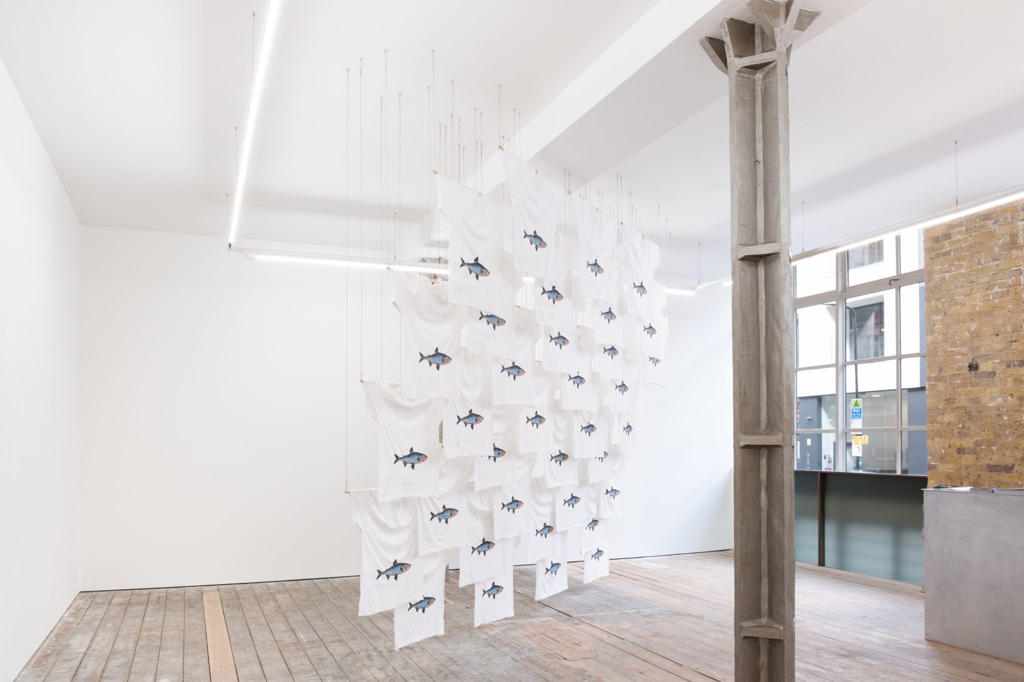 An art installation featuring white fabric panels with images of fish, suspended in a bright, minimalist gallery space with wooden flooring and concrete columns.