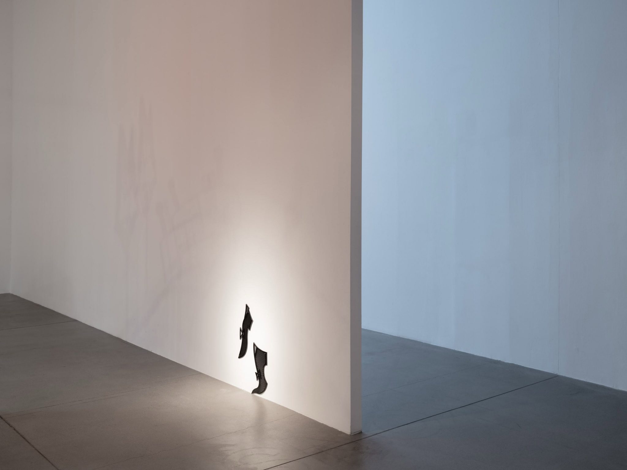 An installation featuring a wall with a shadow cast by an object protruding from it, creating an interplay of light and shadow in a minimalist space.