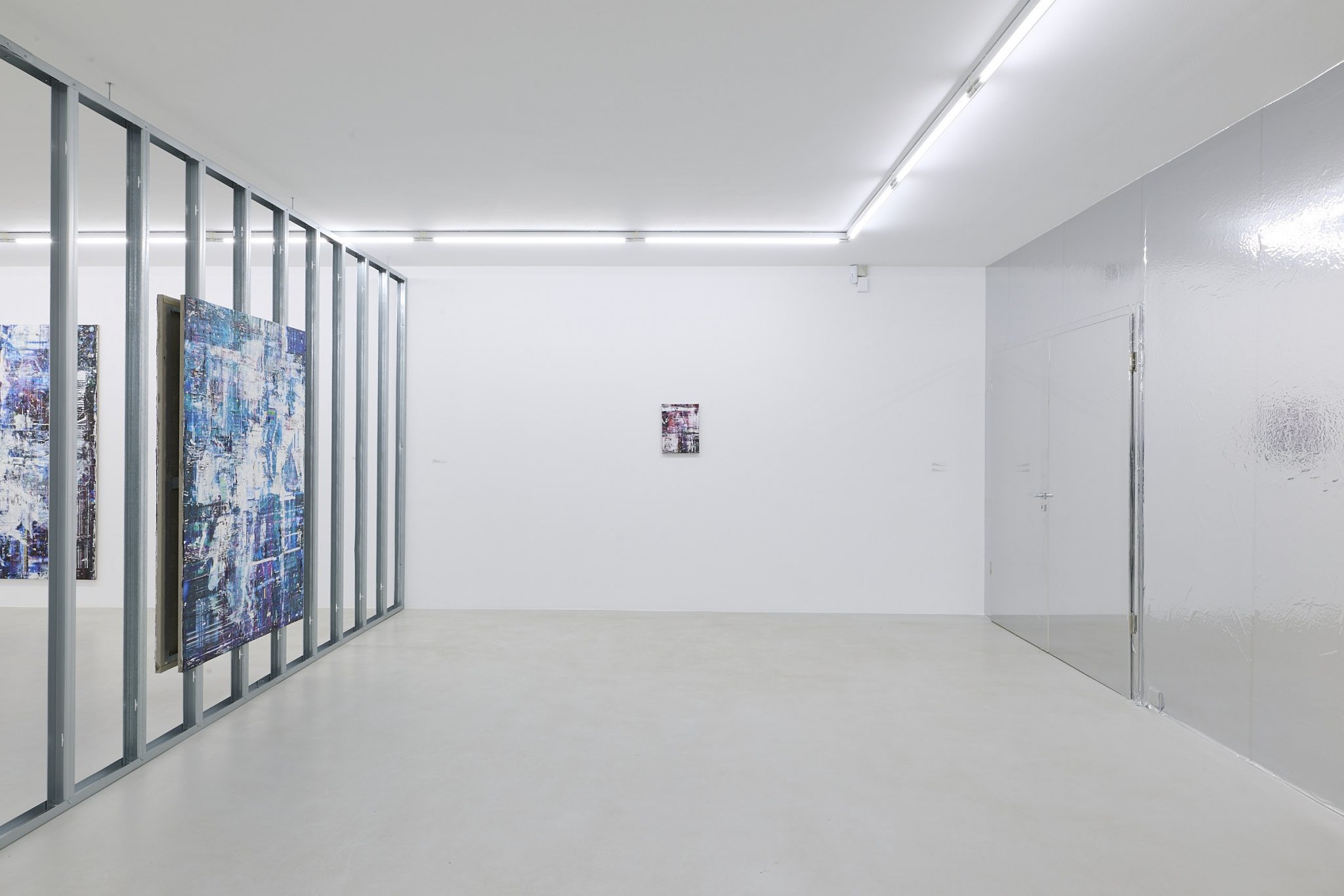 An interior view of a modern art gallery featuring a minimalistic design, with large, light-colored walls and floors. There are abstract paintings on the walls, and a shiny, reflective wall section on one side. The space is well-lit with fluorescent lighting.