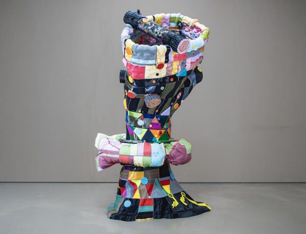A tall, abstract sculpture made of colorful fabric and various materials, resembling a tree-like form with a prominent bulbous top and intricate patterns throughout.
