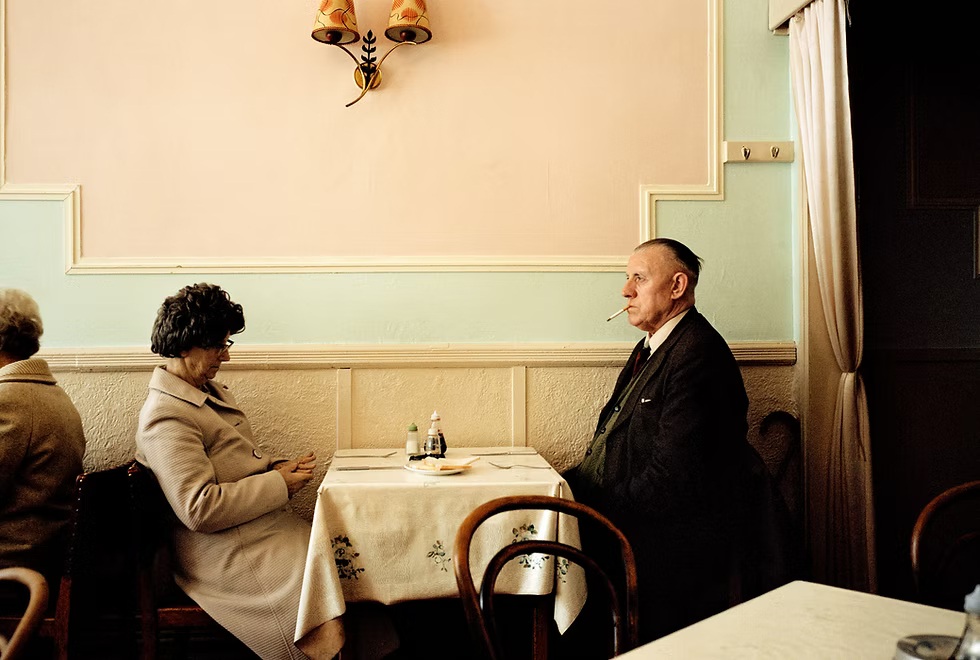 In a warmly lit vintage restaurant, a woman in a beige coat faces an older man in a dark suit, creating a quiet, formal mood.