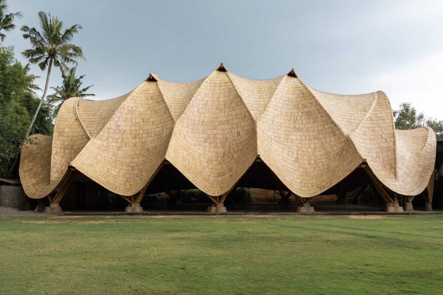 Traditional tropical pavilion with a dramatic woven thatched roof, open sides, palm trees, and a broad green lawn under a calm sky.