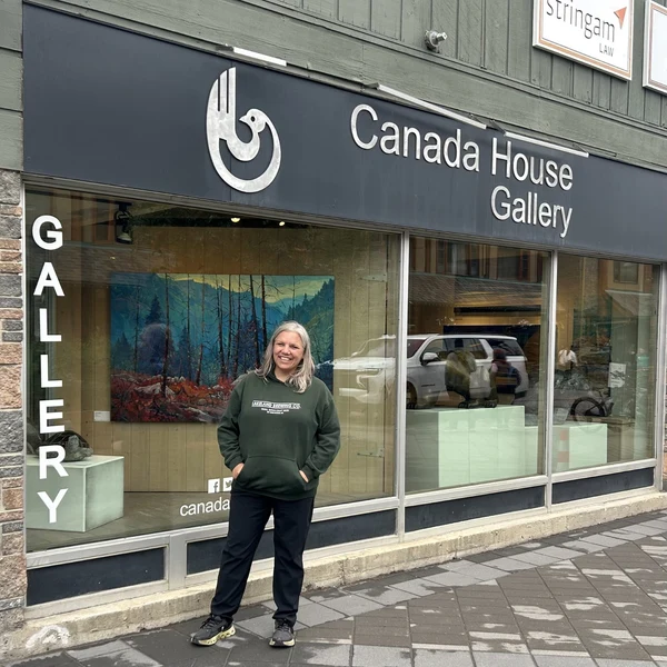 A smiling woman stands outside Canada House Gallery beneath its dark sign, with colorful landscape art in the window; a calm street scene.