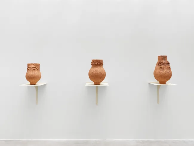 Three small terracotta vessels sit on separate white wall shelves in a minimalist gallery, their varied shapes and carved details creating a quiet, contemplative display.
